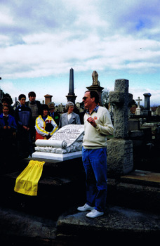 People in a graveyard listening to a man who is standing next to a grave. The grave has a headstone, in the shape of a scroll, mounted on a stack of three stone slabs. At the base of the headstone is a small yellow veil.