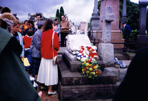 People standing among gravestones in a graveyard. Some are reading the inscription on a headstone, in the shape of a scroll, on a stack of three stone slabs. Words are inscribed on the headstone. Two wreaths of fresh flowers have been placed on the grave.
