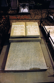 A headstone in the shape of an open book, with inscriptions on the pages, on top of a flat stone slab. 