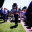 Soldier, head bowed, stands in front of gravestone. Figures look on.