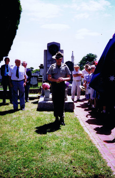 Soldier, head bowed, stands in front of gravestone. Figures look on.
