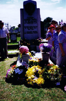 Gravestone covered in floral wreaths. Figures stand nearby.