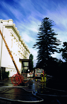 A fire hose and a fire ladder are propped against the front of the white Town Hall building. In the background two fire fighters stand by a fire engine