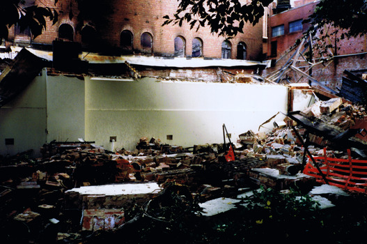 Rubble including bricks, steel girders from a collapsed roof, and burnt furniture and other indistinguishable items, are piled in front of parts of the building that are still standing. 