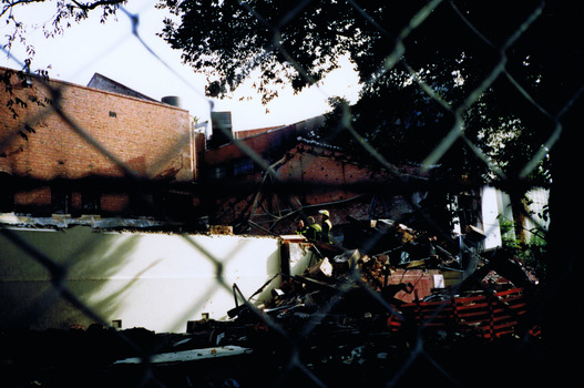 Two fire fighters are seen through a wire fence stand. They stand next to a partially collapsed white wall and bricks, broken furniture and other rubble.