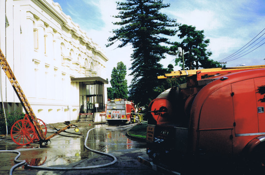 A fire hose and a fire ladder are propped against the front of the white Town Hall building. In the foreground is the rear of a fire engine. Another fire engine is in the background, parked at the entrance to the building, where two fire fighters are working.
