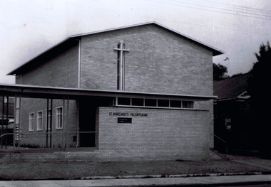 Black and white image of light coloured brick building. On the front is a large cross, above a verandah that extends to the left of the entrance. On the front brick wall a sign reads St Margaret's Presbyterian