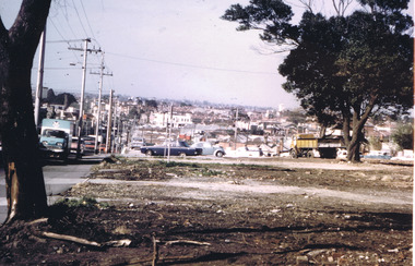 A vacant plot of land after demolition of building. A flat bed truck to remove rubble is parked by a tree on the right. A truck and cars in traffic are on the left.