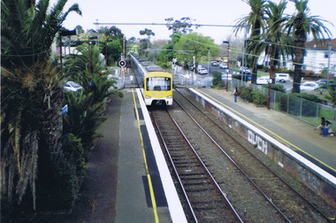 A yellow and silver electric train is crossing a railway crossing as it approaches the station from the city. In the foreground are station platforms on either side of the track. No one is waiting on the platform for the arriving train, Three people are waiting on the platform for a train to the city.