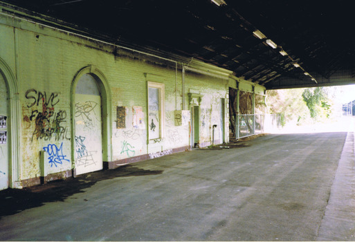An empty, disused, station platform, covered by a wooden roof. The brick walls of the station building are painted with graffiti.