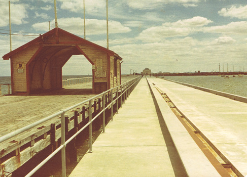On the left is a wooden archway which is at the entrance to a wooden pier. On the right, a concrete pier runs parallel to the wooden pier and leads to a building at the end, which is the St Kilda kiosk.  