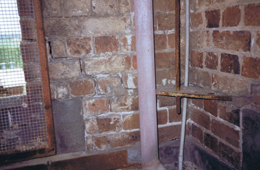 Pipes attached to the inside of a brick building near a window that is covered with rusty wire mesh
