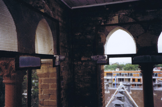 Buildings seen from a height through arched stone and brick windows covered in rusty wire mesh