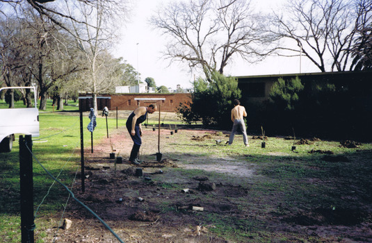 Two men are planting trees and shrubs. One is standing side-on, using his foot to tamp the ground next to a newly planted shrub. The second man is digging with a shovel and is seen from behind. Black plastic pots containing shrubs and trees ready for planting are placed around the area. 