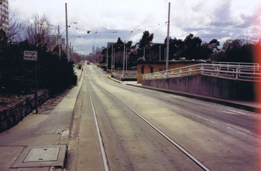 A set of tram tracks extending in a straight line. A second set of track joins them on the right. In the distance is the skyline of Melbourne city. A sign on the left in the foreground says 'pedestrians prohibited'. 