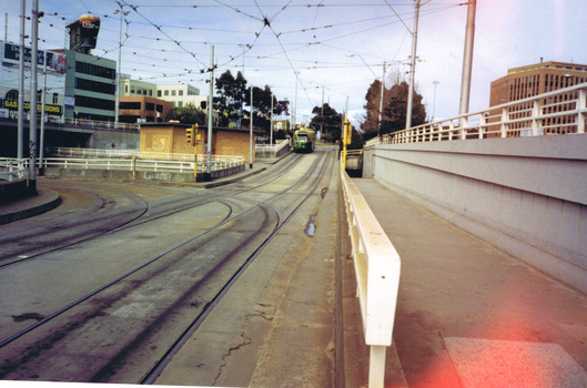 Two sets of tram tracks. One set of diverts to the left and disappears from view under an overpass. The other set extends in a straight line, slightly uphill. A green and yellow tram is on the left, travelling up the hill. On the right is a separated pedestrian walkway that extends downwards in the distance to join a subway under St Kilda junction.
