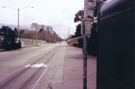 A set of tram tracks extending in a straight line. On the left, two women wait at a green wooden shelter at a tram stop to the city. On the right is a partial view of the sign for a tram stop for routes 5 and 64. 