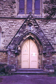 Double solid wood doors, painted yellow, shaped at the top to fit a pointed arch doorway. On the lower half of each door are two recessed rectangular panels, one above the other. On the upper half is a large panel in the shape of a pointed arch and a small panel triangular panel.