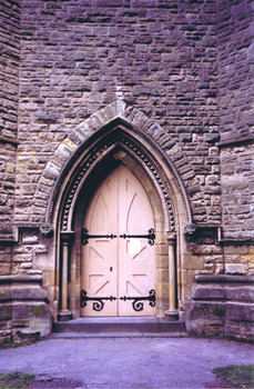 Double solid wood doors, painted yellow and shaped at the top to fit a pointed arch entrance. Each door has two large ornate black hinges, the width of the door, placed at the intermediate and bottom rails. On the lower half of each door are four recessed triangular panels. On the upper half is a large panel in the shape of the arch.