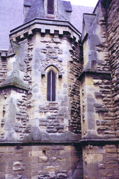 Multi-angular sandstone tower with a pointed roof supported by buttresses. A narrow pointed arch window is placed half way up the tower. 