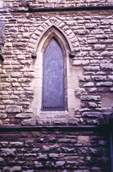 A pointed arch window set in the sandstone wall, left of the northern transept entrance. The image in the stained glass window is obscured by wire mesh. There are stones missing from the wall and other signs of water damage and weathering.