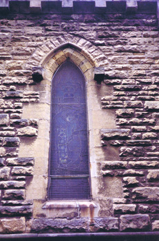 A pointed arch window set in the sandstone wall, right of the northern transept entrance. The image in the stained glass window is obscured by wire mesh