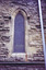A pointed arch window set in the sandstone wall, to the right of the northern transept entrance. The image in the stained glass window is obscured by wire mesh. Metal drainpipes have been attached to the stone wall below and to the right of the window.