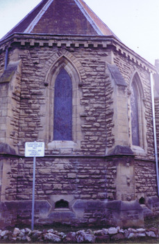 Two pointed arch windows, in two of the three stone walls that form the apse of the church. A supporting buttress is on each side of each window. Next to each buttress is a vertical metal drainpipe. A metal pipe also runs horizontally under the windows. A sign on a metal post in front of the apse says 'Playground for young children only. Football and cricket prohibited.' 