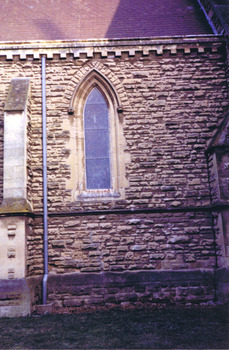 A pointed arch window set in the sandstone wall. A buttress is to the left of the window. Between the window and the buttress is a metal drainpipe.