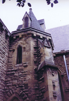 Multi-angular sandstone tower with a pointed tiled roof supported by buttresses. A narrow pointed arch window is placed half way up the tower. 