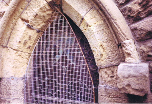 The apex of a pointed arch window of stained glass that is obscured by wire mesh. The stonework around the window is eroded. 