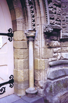 The right hand side door of double solid wood doors, painted yellow and shaped at the top to fit a pointed arch entrance. The door has two large ornate black hinges, the width of the door, placed at the intermediate and bottom rails. Adjacent to the archway entrance, on the right, is a sandstone colonette.
