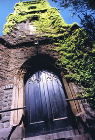 A solid wooden door, painted black, in a gothic pointed archway of sandstone at the entrance to a bluestone tower. Above the doorway is a pointed arch window and, above that, a round window. The very top of the tower cannot be seen. Bluestone steps lead up to the door. Ivy grows up the full height of the tower. 