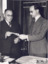 Black and white photograph showing Sir Frank Beaurepaire and Mr WH Greaves standing in front of a desk and examining papers being passed between them