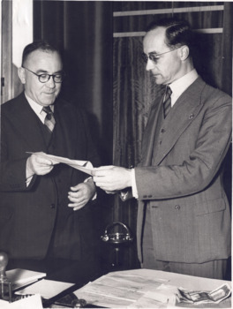 Black and white photograph showing Sir Frank Beaurepaire and Mr WH Greaves standing in front of a desk and examining papers being passed between them
