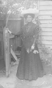 Photograph, Unknown young Lady dressed with hat and gloves standing beside a house gate c1910