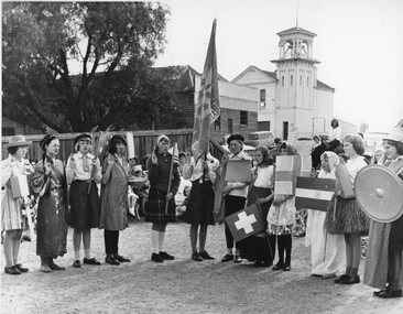 Photograph, Girl guides in dress up with flag, at guide hall with blacksmith shop and fire station in background