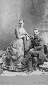 Photograph, Carte-de-Visite Studio photo of Gentleman seated with lady in multi layered dress standing.  c1870