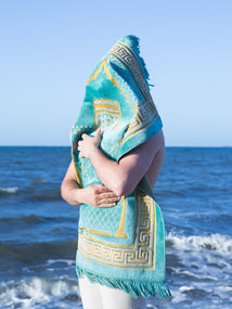 This photograph was the hero image from artist Ayman Kaake's commissioned exhibition at Eltham Library Community Gallery 'In their names' which was Nillumbik Shire's first collaboration with Midsumma Festival.  The work is a self-portrait of the artist at Half Moon Bay in which he is covered by and holds a Prayer Rug.  The back drop is the sea, a significant border between countries in the artists work.