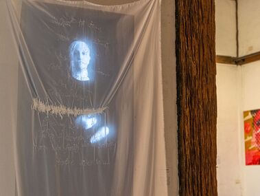 White sheer fabric embroidered with text and sound waves is suspended in front of a black and white video work, portrait aspect ratio, that shows the artist signing