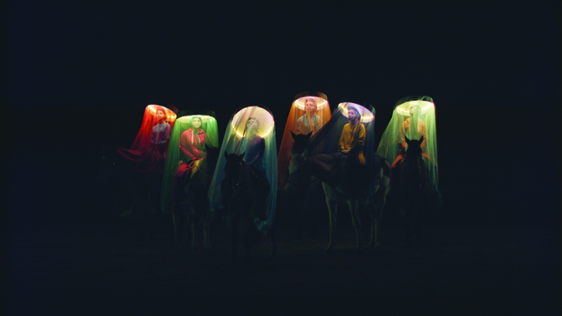 6 women sit mounted on horses in a darkened space.  They each wear a head piece with a circular light around the head that is covered with a gauze like veil that comes down to the horses legs and lights their faces. Each is a different colour - red, green, blue, orange, purple, cream.