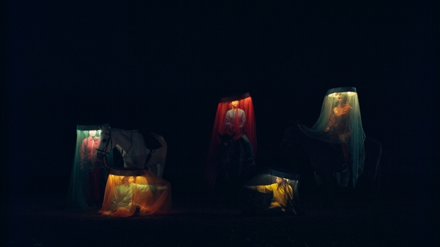 6 women sit mounted at different heights on horses in a darkened space.  They each wear a head piece with a circular light around the head that is covered with a gauze like veil that comes down to the horses legs and lights their faces. Each is a different colour - red, green, blue, orange, purple, cream. Two of the riders point to each other.