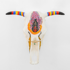 A 3D printed bull skull with horns painted in vibrant blue, red and  yellow stripes and black painted tips, and a redback spider painted on the centre of the skull.  A small crucifix is attached to the redback spider and beaded jewellery is looped through the eyes of the skull.
