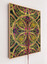 The work has a symmetrical print with green and red coloured leaf shapes featured as its base. There are elements of embroidery of yellow wattle like flowers stitched onto the surface of the prints. There is bright pink stitching on the surface as well. Coloured floss twine is also attached on the surface of the work that hangs down beyond the dimension of the frame at the bottom of the work