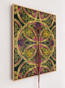 The work has a symmetrical print with green and red coloured leaf shapes featured as its base. There are elements of embroidery of yellow wattle like flowers stitched onto the surface of the prints. There is bright pink stitching on the surface as well. Coloured floss twine is also attached on the surface of the work that hangs down beyond the dimension of the frame at the bottom of the work