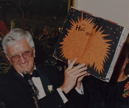 A large book open to an artwork page being held up by a man standing at a microphone.