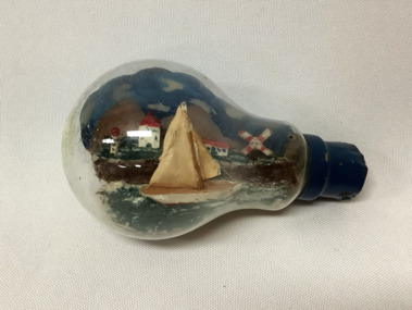A small model sailing boat in a glass light bulb, with a lighthouse, a windmill and three buildings on a cliff top in the background.