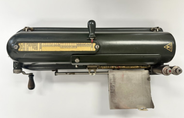 A vintage machine for printing cash amounts on cheques. The cheques are rolled through the machine by placing them on the flat metal platform and then turning the handle to print a preset amount. Has a black vinyl cover. 