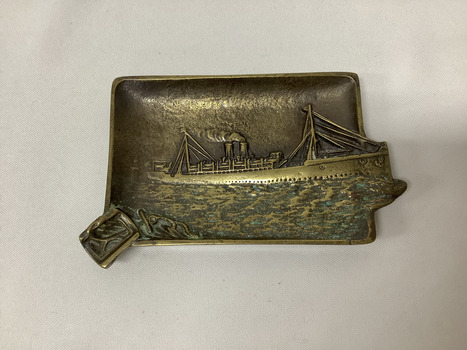 A rectangular brass ashtray depicting, in relief, the paddle steamer PS Hygeia. There is a groove in the bottom left hand corner for cigarette placement.