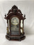 Eight-day mantle clock in an ornate carved wooden case. Plain face with Roman numerals,  under glass. A glass door allows access to the weighted pendulum, and is decocrated with an etching-like transfer showing a bearded Cavalier with a sword. The clock face and pendulum is flanked by a pair of brass or gold coloured cupids, standing on turned wooden pedastels, with mirrors behind them. A carved wooden head is above the face, and another carved wooden head flanked with wings is on the base.
